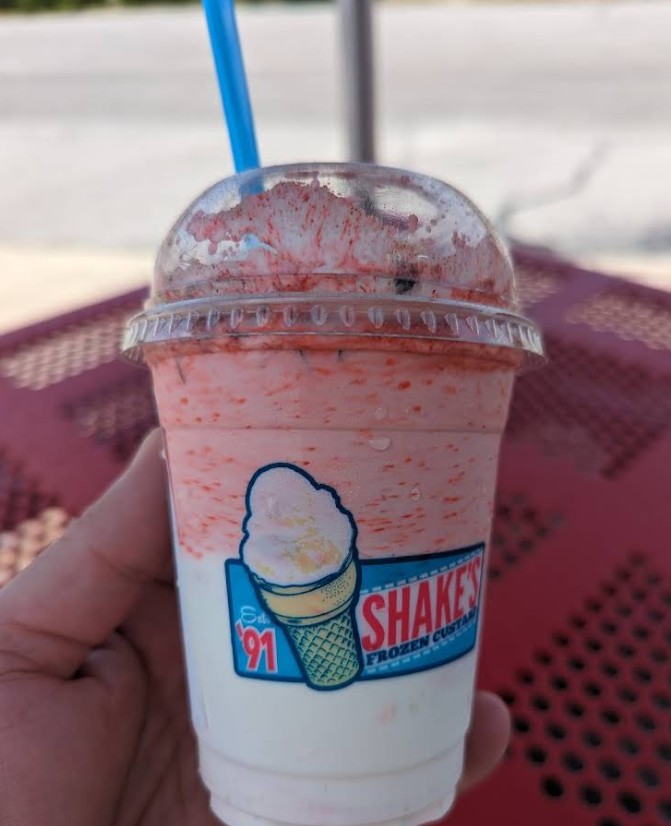 shakes | Beach Condos in Destin The Best Ice Cream in Miramar Beach and Destin #Strawberry milkshake in a clear cup with a blue straw, held outdoors, with red strawberry sauce on top and sides.