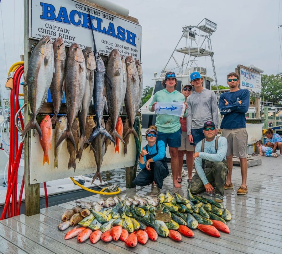 Watersport Activities in Destin | Beach Condos in Destin #Group of anglers on a dock posing with dozens of large fish hanging on a rack beside a'Backlash' charter boat sign.