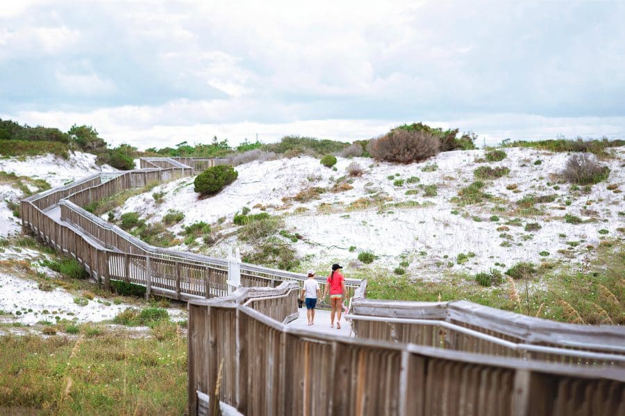State Parks Near Destin/Miramar Beach | Beach Condos in Destin #Wooden boardwalk winding through white sand dunes with shrubs, two people walking away under a cloudy sky.