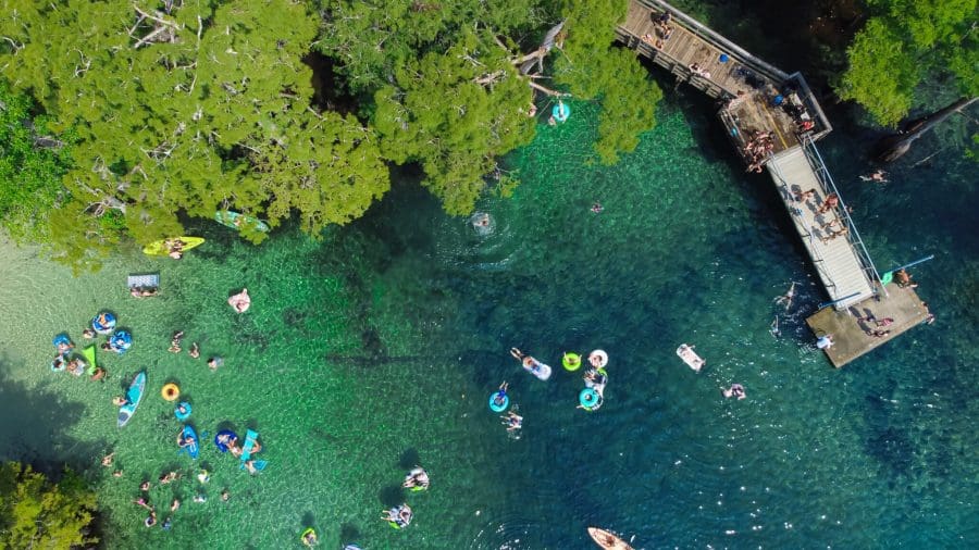 Crowded of people enjoy floating swimming kayaking jumping of | Beach Condos in Destin Cool Off at Florida’s Natural Springs Near Destin | Beach Condos in Destin #Aerial view of a sunny lake with turquoise water, people swimming and floating near a wooden dock by lush green trees.