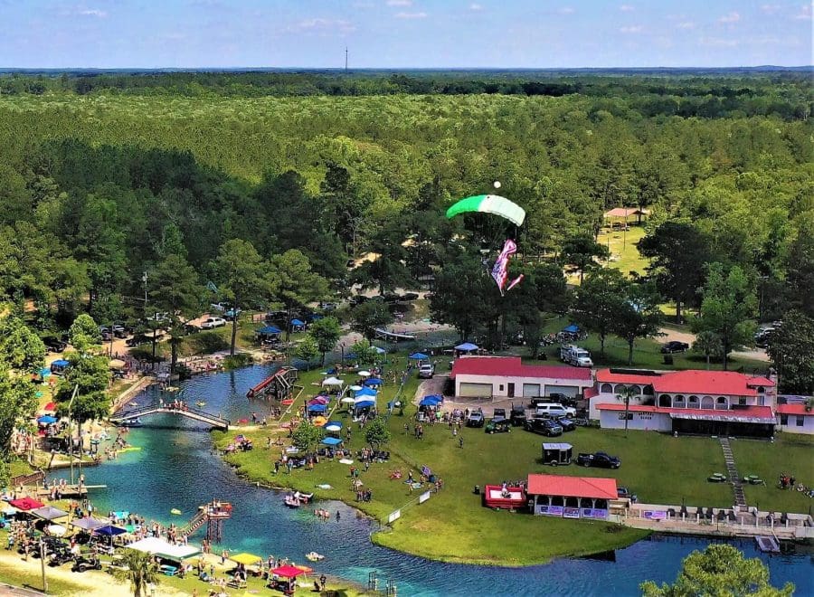 Vortex-Spring-Florida-2026 | Beach Condos in Destin Cool Off at Florida’s Natural Springs Near Destin | Beach Condos in Destin #Aerial view of a lakeside festival with tents, people, and a green-and-white paraglider in flight above the crowd.
