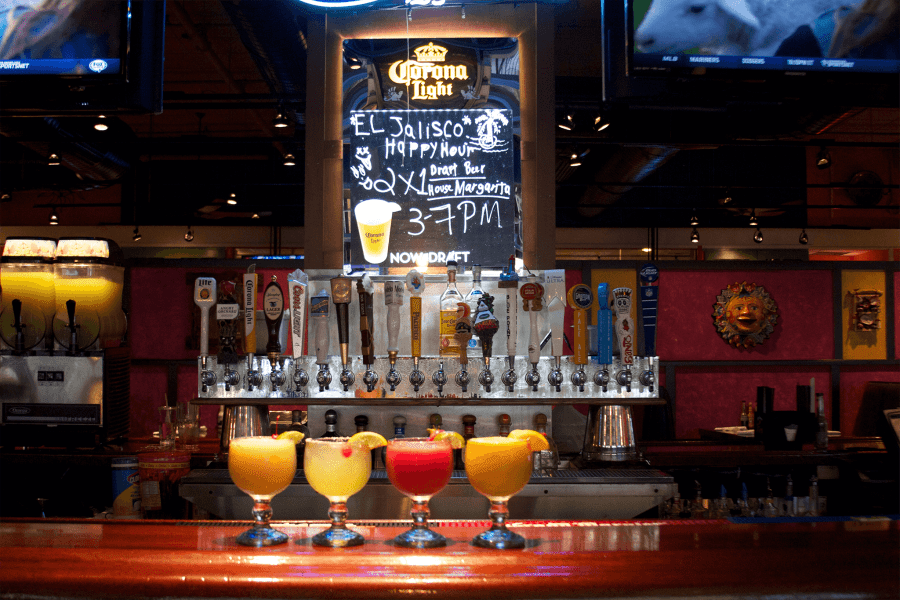 Bar scene with beer taps in the background, a Corona Light sign, a chalkboard menu, and four colorful cocktails in the foreground.
