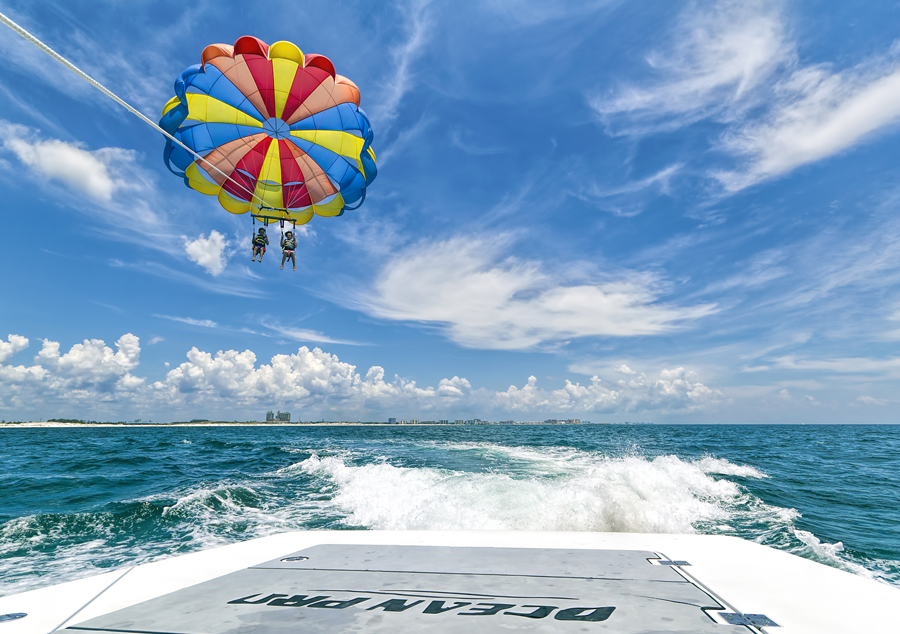 Watersport Activities in Destin | Beach Condos in Destin #Parasailers ride a bright multicolor parachute over blue sea, tethered to a speeding boat.