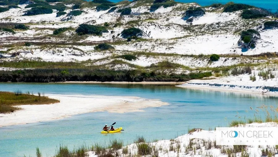 State Parks Near Destin/Miramar Beach | Beach Condos in Destin #Two people kayaking on a shallow turquoise river with white sandy banks and dunes in the background.