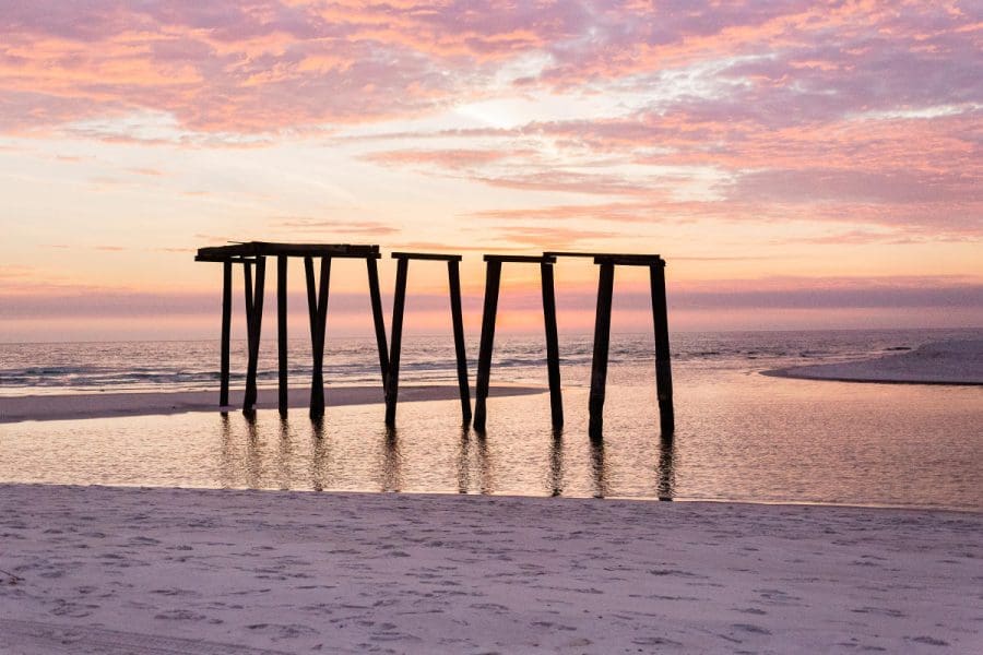 State Parks Near Destin/Miramar Beach | Beach Condos in Destin #Sunset over a calm sea with a weathered wooden jetty extending into the water, silhouetted by pink-orange skies.