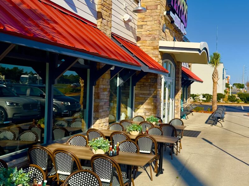 Outdoor cafe seating along a storefront with red awnings and stone facade under a clear blue sky.