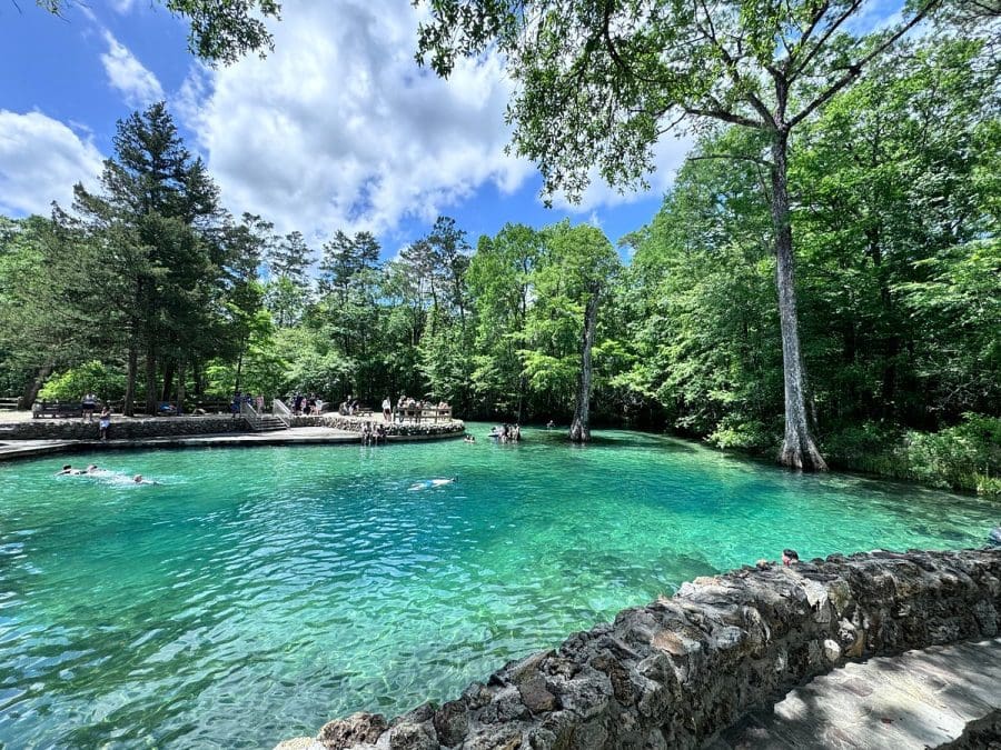 ponce de leon spring 2026 | Beach Condos in Destin Cool Off at Florida’s Natural Springs Near Destin | Beach Condos in Destin #Turquoise natural pool surrounded by a forest with people swimming and a stone railing along the edge.