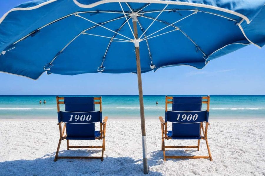 1900-ninety-eight-beach-servicejpg_1535w | Beach Condos in Destin 1900 Ninety Eight | Beach Condos in Destin #Two blue beach chairs with '1900' on the backrests sit under a large blue umbrella on white sand, with the turquoise ocean in the background and a clear sky.
