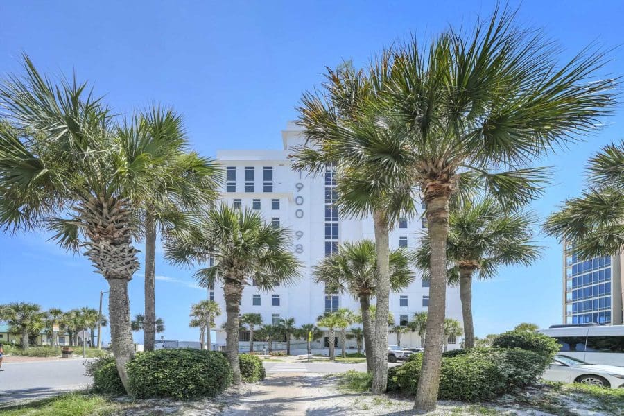 1900-ninety-eight-destinjpg_1920w | Beach Condos in Destin 1900 Ninety Eight | Beach Condos in Destin #White mid-rise building seen behind several palm trees on a sunny day with a clear blue sky.