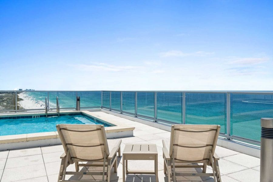 1900-ninety-eight-rooftop-pooljpg_1920w | Beach Condos in Destin 1900 Ninety Eight | Beach Condos in Destin #Rooftop pool area with beige lounge chairs and a glass railing overlooking the blue ocean under a clear sky.
