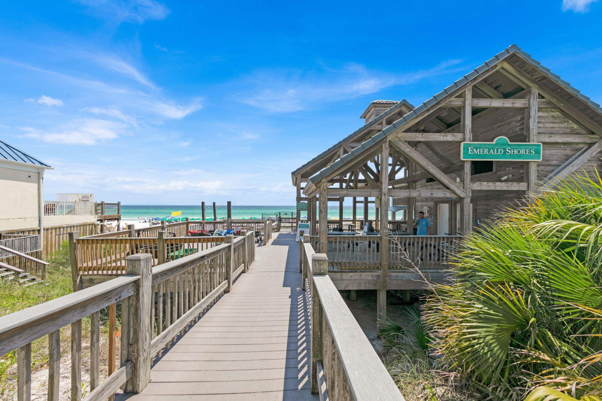 Beach Condos in Destin FL | Vacation Rentals Online #Beachfront wooden boardwalk leading to a rustic pavilion with an Emerald Shores sign, blue sky and ocean beyond