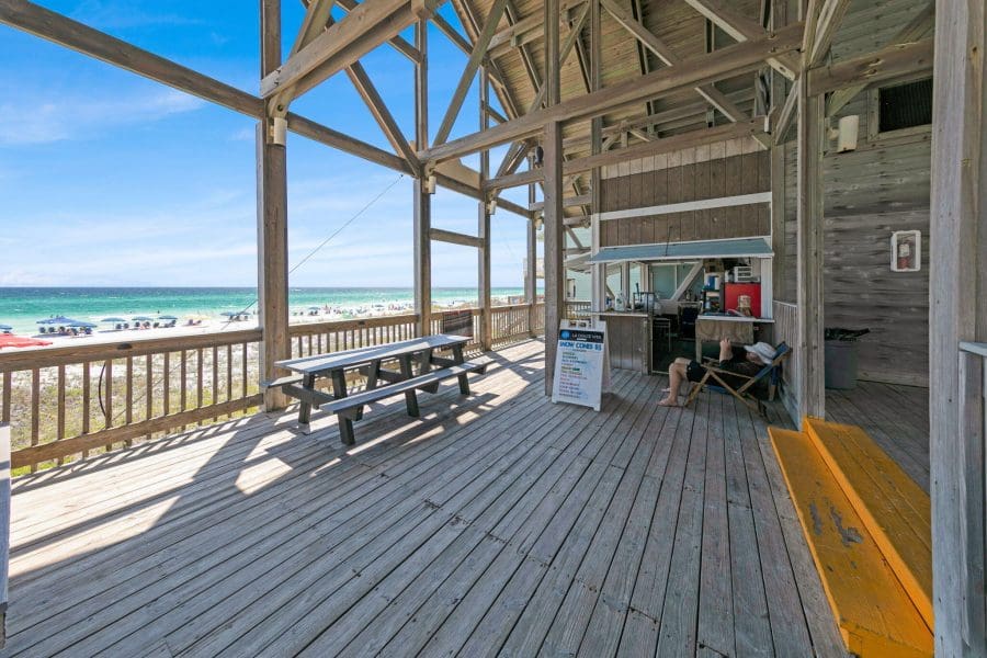 Emerald Shores | Beach Condos in Destin #Wooden beach boardwalk with picnic table under a wooden structure, overlooking a sunny beach and turquoise sea with umbrellas in the sand
