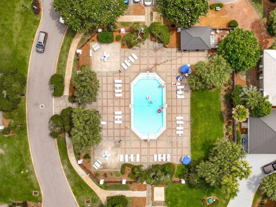 Emerald Shores | Beach Condos in Destin #Aerial view of a rectangular turquoise swimming pool surrounded by white lounge chairs, blue umbrellas, and palm trees in a tiled patio area within a landscaped resort yard.
