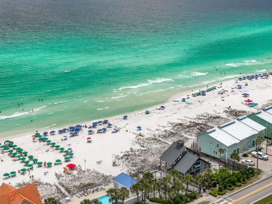 Emerald Shores | Beach Condos in Destin #Aerial view of a crowded white-sand beach with turquoise water and numerous colorful umbrellas, adjacent to beach houses and a road.