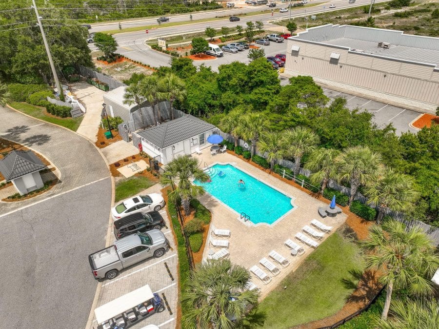 Emerald Shores | Beach Condos in Destin #Aerial view of a pool area with lounge chairs and palm trees behind a beige building, with a parking lot and road with traffic beyond.