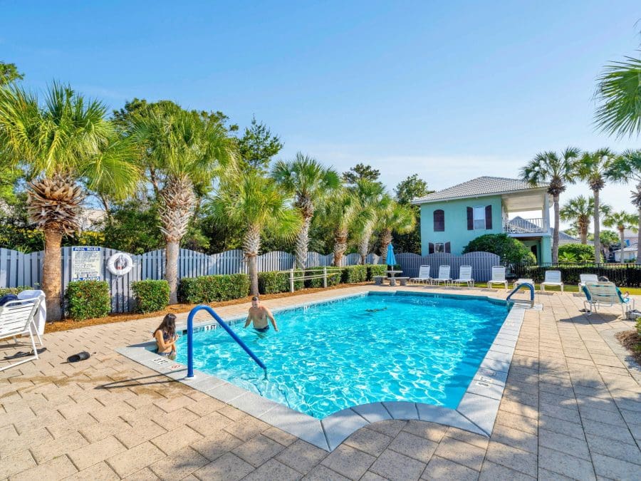 Emerald Shores | Beach Condos in Destin #Pool with people swimming, surrounded by palm trees and lounge chairs under a bright blue sky