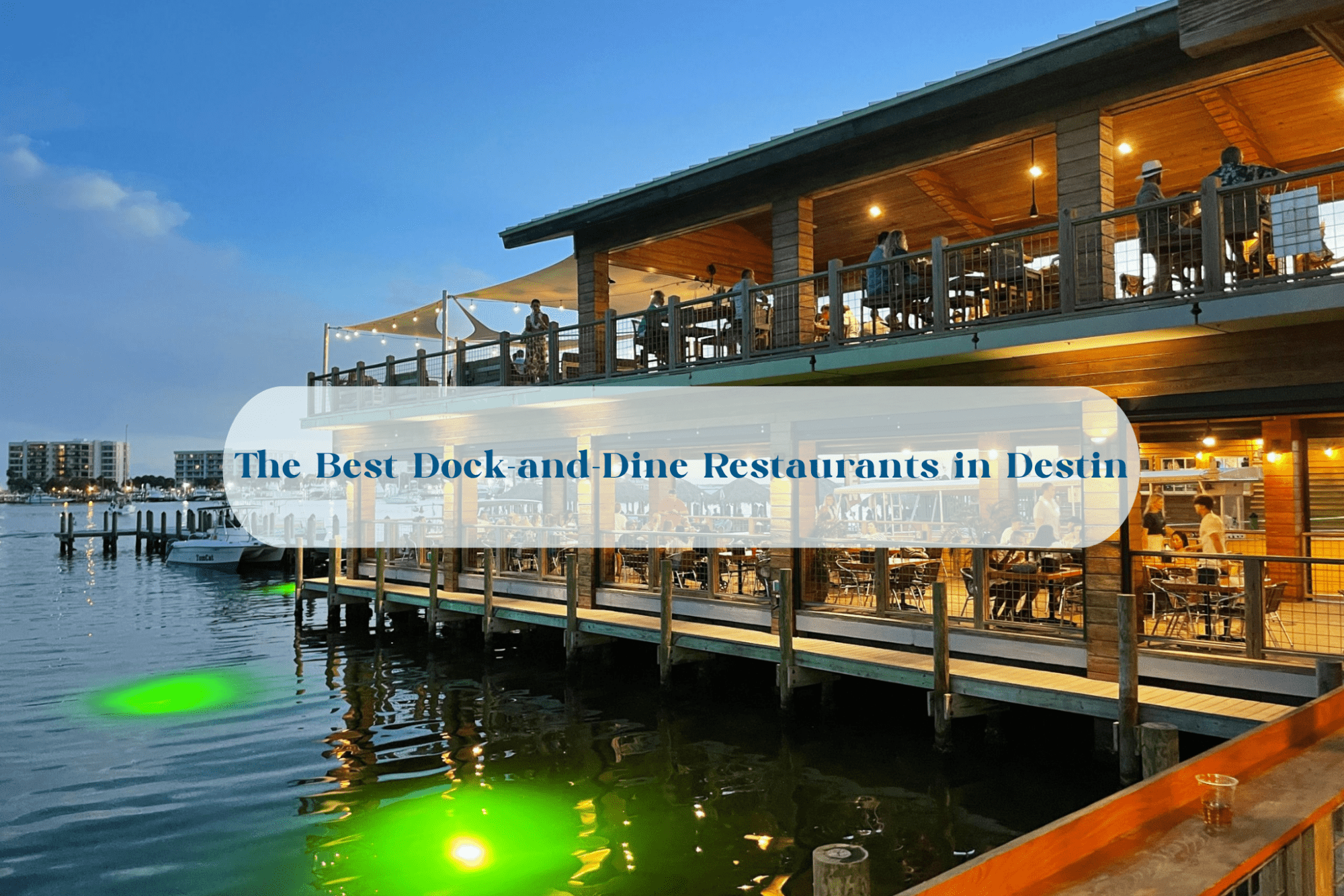 Boat-Up Restaurants in Destin FL | Beach Condos in Destin #Two-level dockside restaurant over calm water at dusk, with diners and string lights.