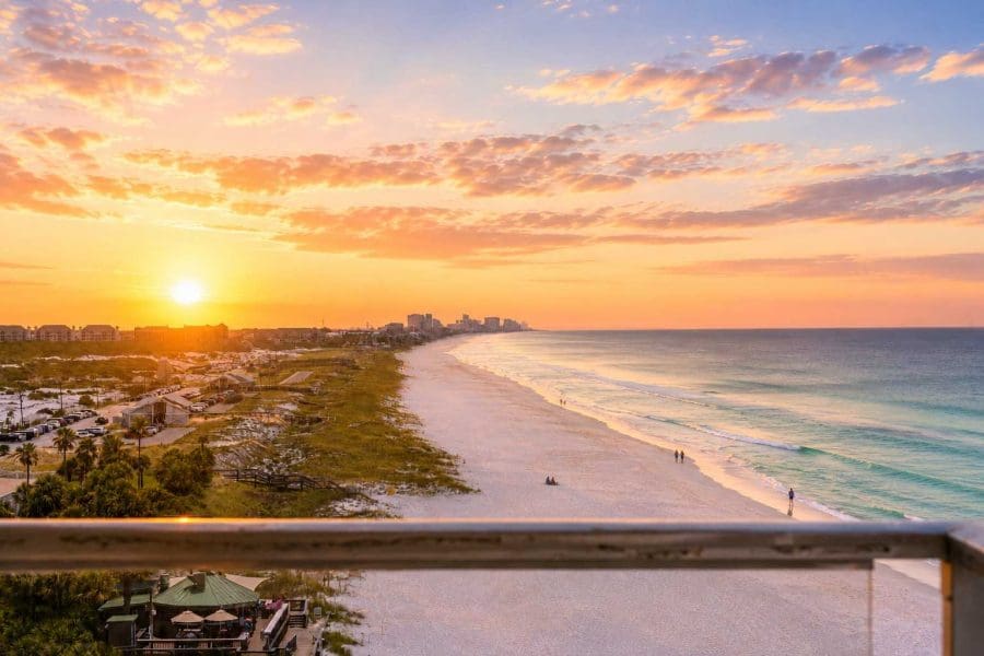 Sunrise Gulf-front Balcony At 1900 Ninety Eight In Destin_1536w | Beach Condos in Destin 1900 Ninety Eight | Beach Condos in Destin #Sunset over a sandy beach with calm turquoise water, buildings in the distance, and a balcony railing in the foreground.