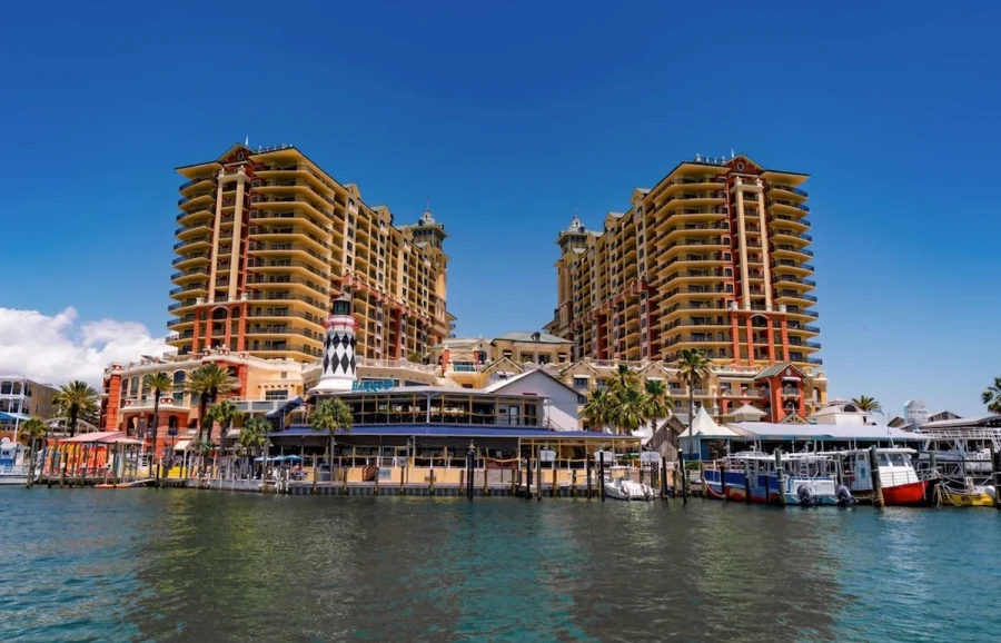 Boat-Up Restaurants in Destin FL | Beach Condos in Destin #Waterfront resort with tall tan and red-brick towers, a marina, and boats docked along a pier under a clear blue sky